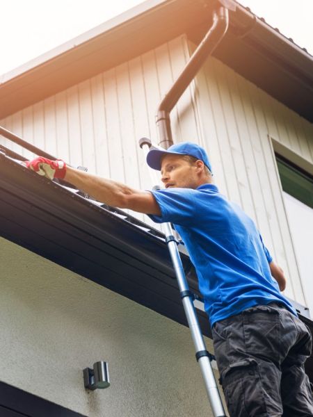 Man on ladder cleans home gutters. Wearing a blue shirt and gloves, he removes debris from the gutter system, ensuring proper water drainage and home maintenance.