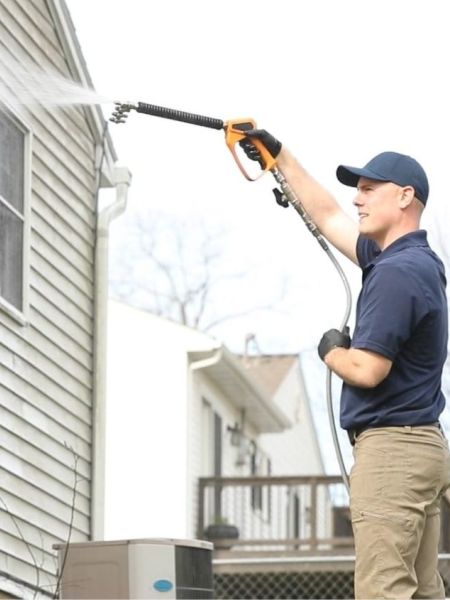 Man cleaning home gutters with a pressure washer extension wand, wearing work clothes and a baseball cap. The house has light siding and a window.
