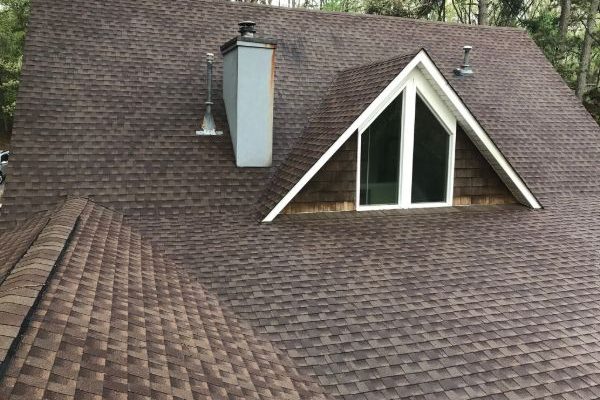 Aerial view of a brown asphalt shingle roof with a chimney and a triangular dormer window. The roof's texture and color variations are visible, suggesting potential need for roof cleaning.