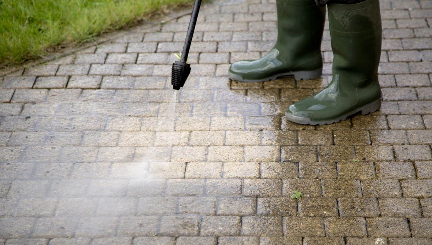 Person in green rubber boots pressure washing a brick paver patio. Half of the patio is clean, demonstrating the effectiveness of paver sealing and cleaning.
