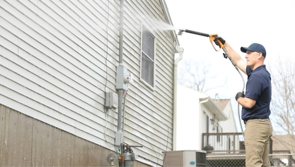 Man power washing the side of a house with siding. He wears a blue shirt and hat, tan pants, and black gloves, holding the pressure washer wand up to clean the exterior. House washing maintenance.