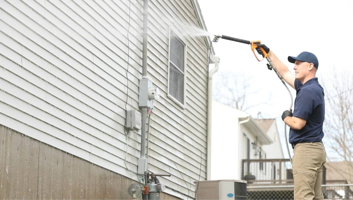 Man power washing the side of a house with siding. He wears a blue shirt and hat, tan pants, and black gloves, holding the pressure washer wand up to clean the exterior. House washing maintenance.