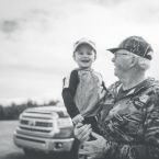 Grandfather in camo holds smiling grandson in ball cap outdoors near a pickup truck. A heartwarming moment, perhaps after gutter cleaning work.