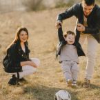 A happy family plays outdoors: a toddler in a black jacket is lifted by his father, while the mother kneels nearby, smiling at the "Home" they've made together.