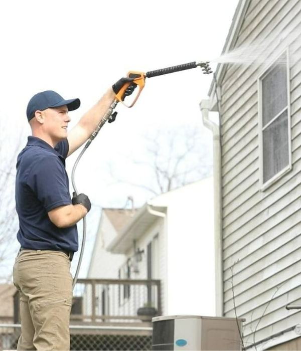 A man power washing the siding of a home. He's wearing a blue cap, gloves, and khaki pants, using a long wand to spray water on the house's exterior. The background shows another house and greenery.