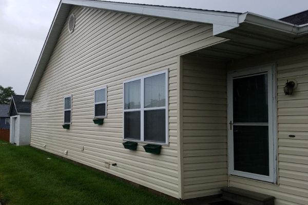 Beige vinyl siding on a house with white trim, windows, and door. Green window boxes add a touch of color. This house washing project showcases clean exterior surfaces.