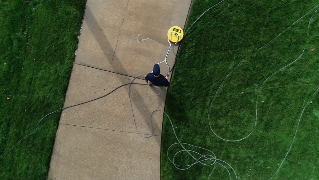 Overhead view of a man pressure washing a concrete walkway with a yellow surface cleaner, surrounded by green grass and a coiled hose. Illustrates a common home maintenance task.