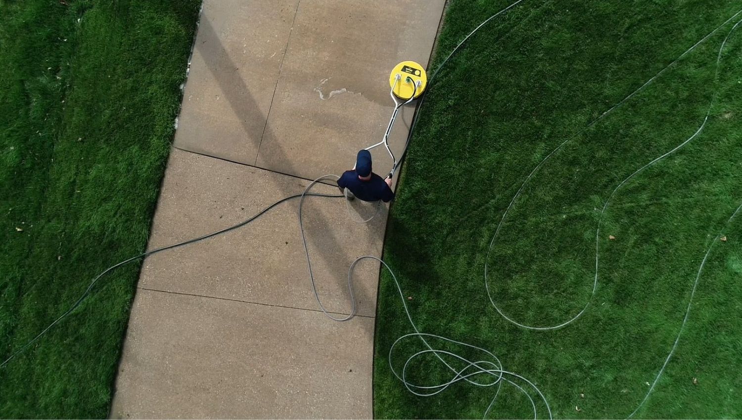 Overhead view of a man pressure washing a concrete walkway with a yellow surface cleaner, surrounded by green grass and a coiled hose. Illustrates a common home maintenance task.