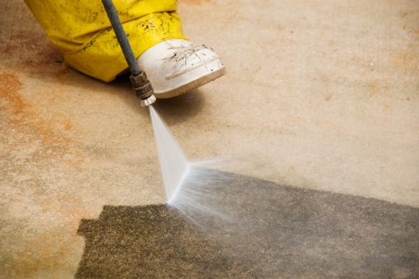 Close-up of a person pressure washing a concrete surface, revealing the clean area underneath. The person wears yellow waterproof pants and white shoes.
