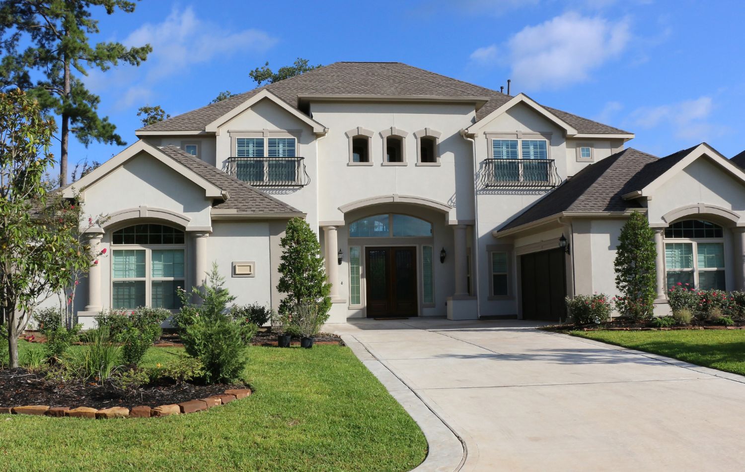 Elegant two-story stucco home with a well-manicured lawn and driveway. The house features a symmetrical design, arched windows, and small balconies, showcasing a beautiful residential property.
