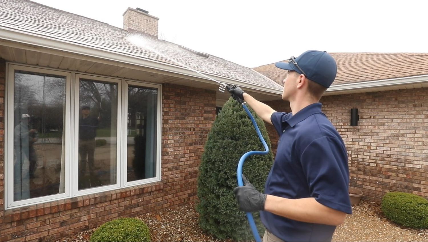 Man power washing a roof with a long wand. Home maintenance and roof cleaning to remove algae and debris. Brick house and large windows visible.