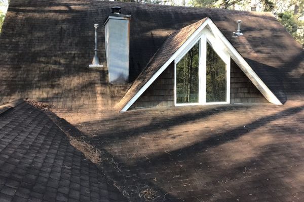 Dark asphalt shingle roof with moss and algae growth, showing a dormer window and chimney. This roof needs cleaning to remove the organic material and restore its appearance.