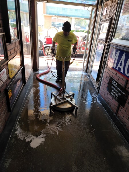 A worker uses a floor cleaning machine on a wet, reflective concrete floor inside a building. A truck with cleaning equipment is visible through the doorway.