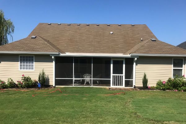 Backyard view of a single-story house with a brown shingle roof and a screened-in porch. Lush green lawn and flower beds add curb appeal.
