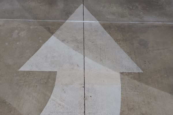 White painted arrow pointing upwards on a weathered concrete surface. The arrow is split by a thin line running vertically, adding a sense of direction and guidance.