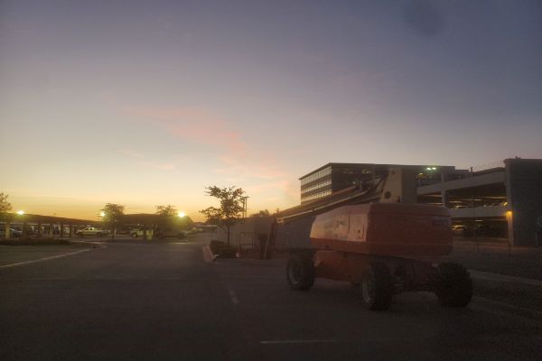 Orange boom lift sits on a paved surface at dusk. The sky fades from pink to blue. A parking garage and office building are in the background.