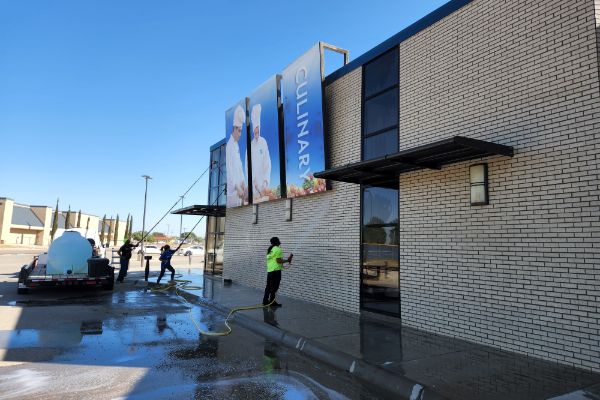 Commercial power washing in progress: Workers clean a brick building with "Culinary" banners using a water tank and long hoses under a clear blue sky.