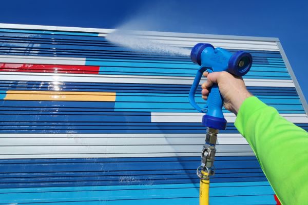 Commercial power washing in action: A hand holds a blue pressure washer, spraying water against a colorful building facade with blue, white, red, and yellow stripes.