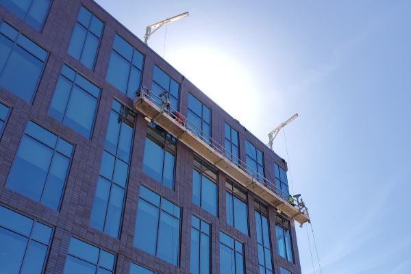 Commercial power washing in progress: Workers on a suspended platform clean the exterior of a modern brick building with large windows, under a bright blue sky.