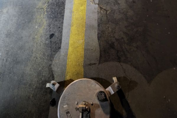 Concrete floor being cleaned with a floor scrubber, revealing a bright yellow line underneath dirt and grime. The cleaning process highlights the contrast between clean and dirty surfaces.