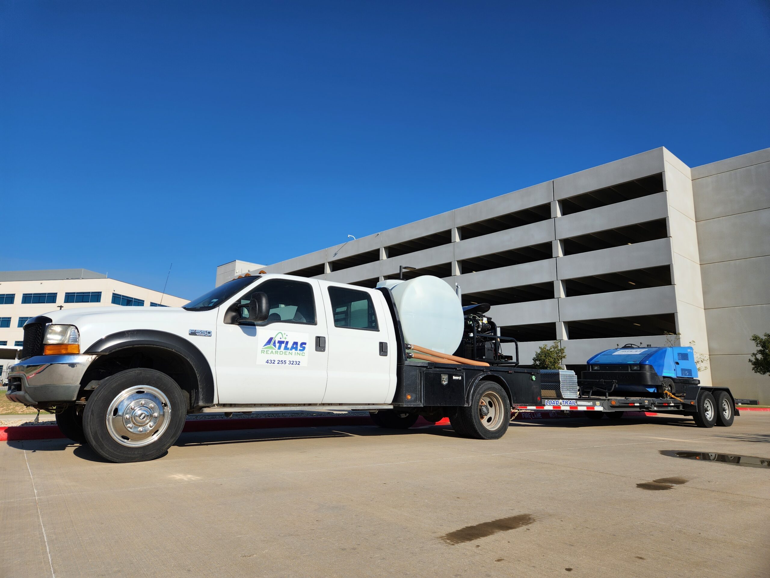 White Atlas Gardening truck with a water tank and trailer equipped for parking lot power sweeping. The truck has "ATLAS GARDENING INC. 432 255 1232" printed on the side. A parking garage is in the background.