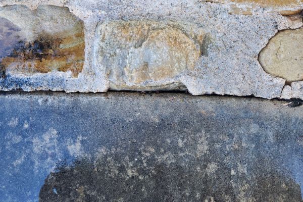 Close-up of a weathered stone wall above a dark, damp concrete surface, possibly after pressure washing. The contrast highlights textures and the effects of moisture.