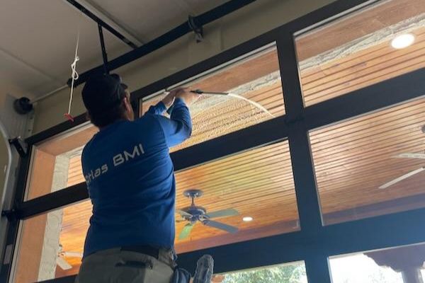 A window cleaner from Atlas BMI uses a squeegee to clean large windows, with a ceiling fan and wood ceiling visible inside the home.