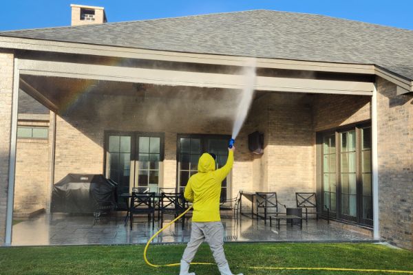 A person in a yellow raincoat power washes a home patio, creating a rainbow in the spray. The house is brick with dark trim, and green grass is visible in the foreground.