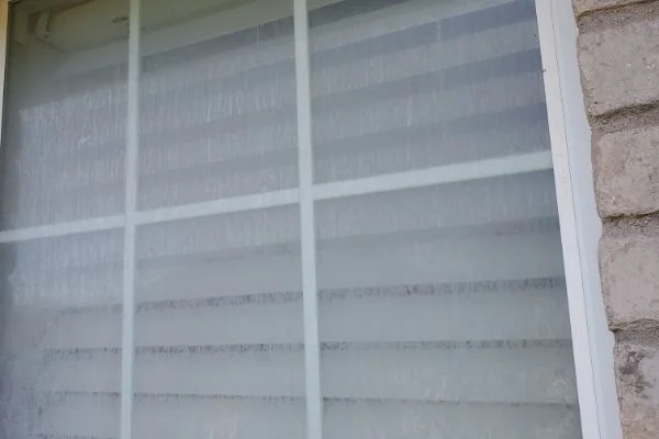 Cloudy glass partially obscures white horizontal blinds behind a window.