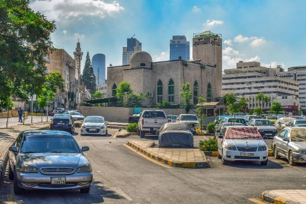 Cars parked in a lot in Amman, Jordan, with a mosque and modern buildings in the background under a partly cloudy sky. Parking lot power sweeping helps keep areas like this clean.