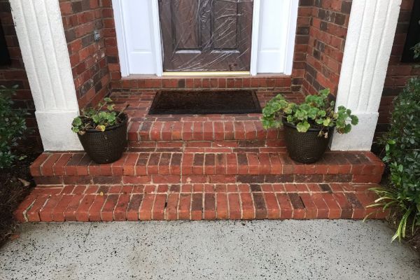 Brick front porch with two steps leading to a brown door covered in plastic, flanked by white columns and potted plants. A dark mat sits in front of the door.