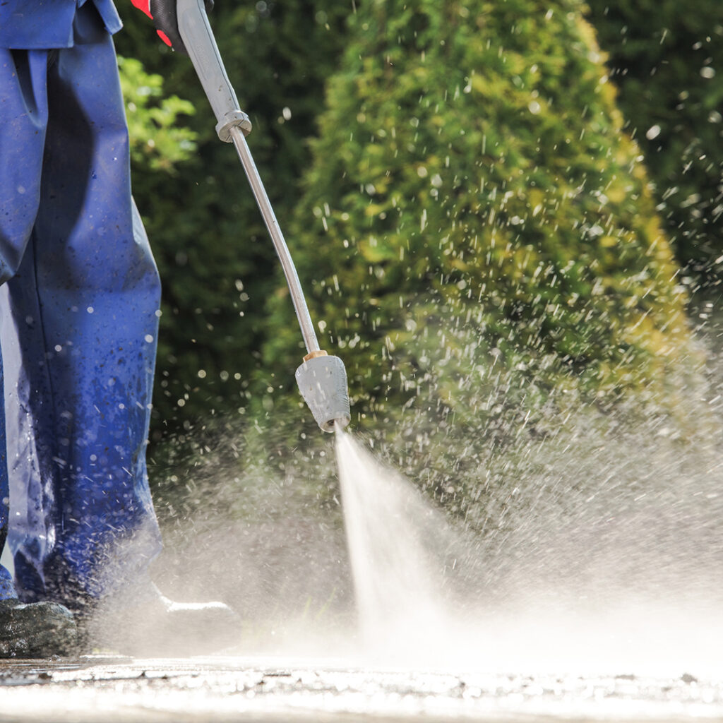 Commercial power washing in action. A worker in blue overalls uses a pressure washer to clean a surface, creating a spray of water droplets against a green backdrop.