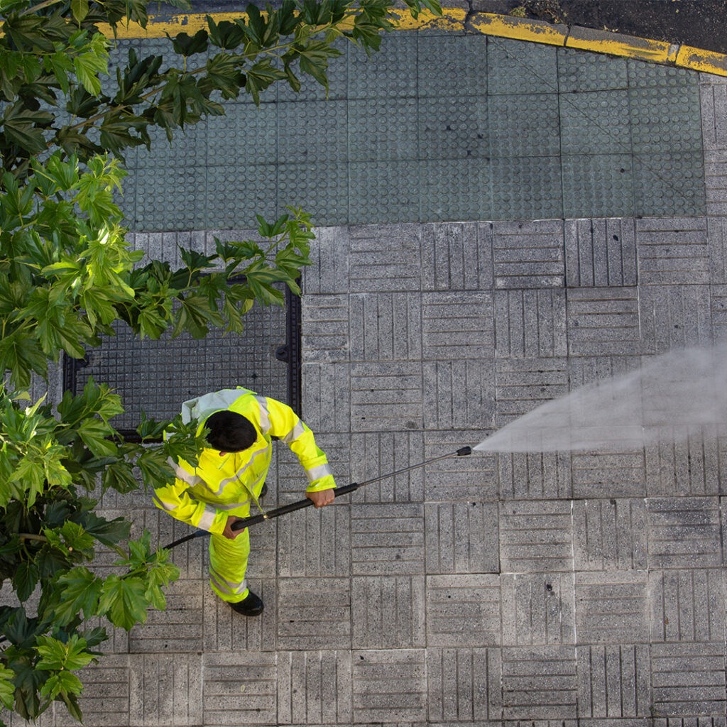 A worker in a bright yellow safety suit uses commercial power washing equipment to clean a grey tiled sidewalk next to a tree-lined street.