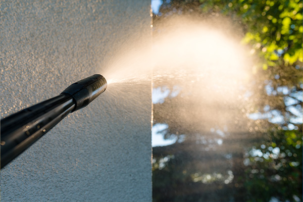 Close-up of a pressure washer spraying a house during house washing. A powerful stream of water cleans the exterior wall, with greenery and trees blurred in the background.