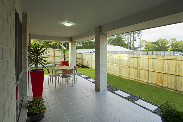Covered patio with gray tile, modern outdoor furniture, and a view of a green lawn and wooden fence. Tall red planters add a pop of color to the scene.
