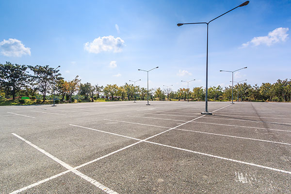 Empty asphalt parking lot with painted white lines under a bright blue sky, surrounded by trees and lit by tall streetlights, ready for power sweeping.