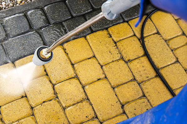 Close-up of a pressure washer cleaning a brick patio. The bricks are yellow and gray, with the pressure washing revealing the clean, vibrant color of the bricks.