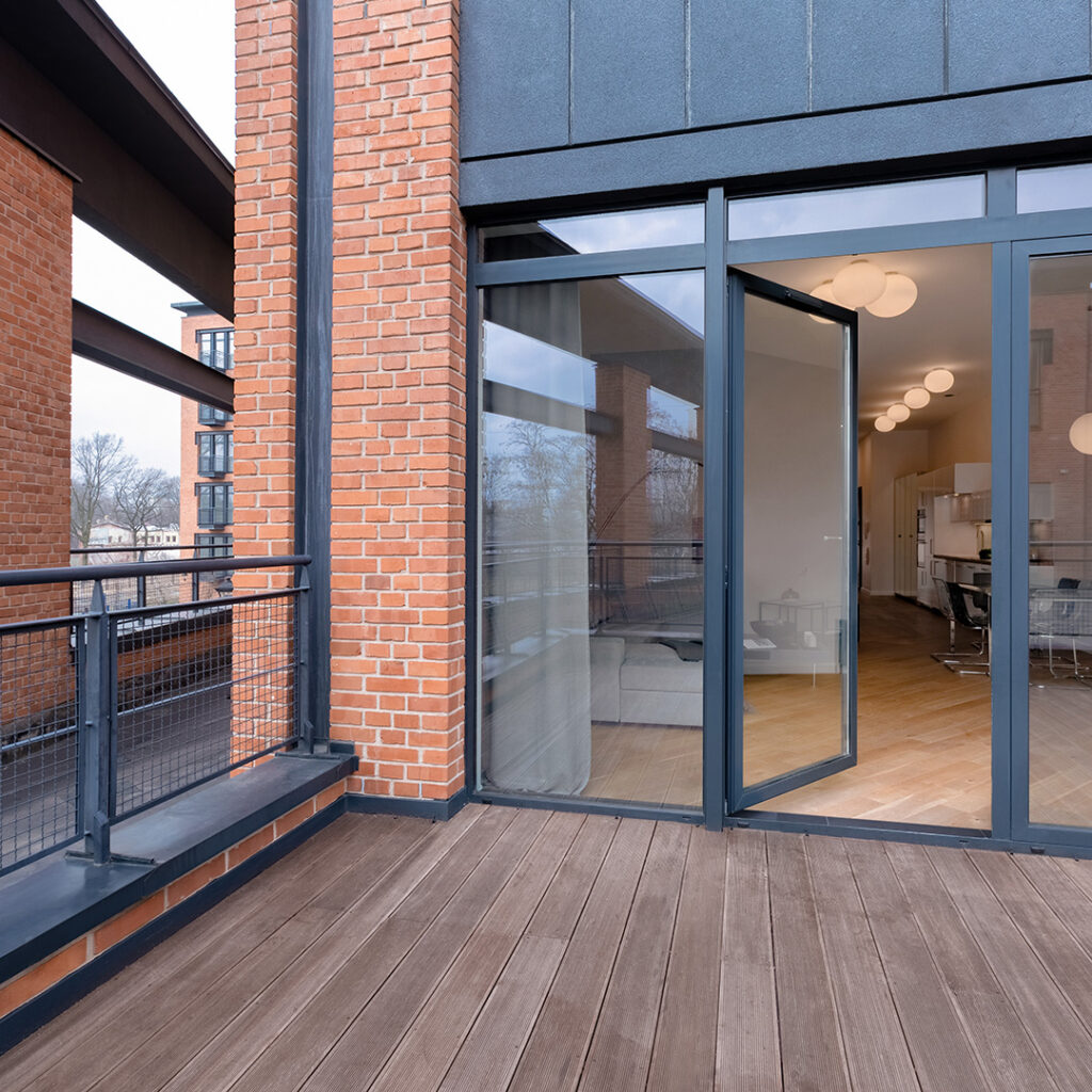 Modern apartment balcony with gray-framed glass doors open to a bright interior. Brick accents, wood deck, and metal railing create an inviting outdoor space.