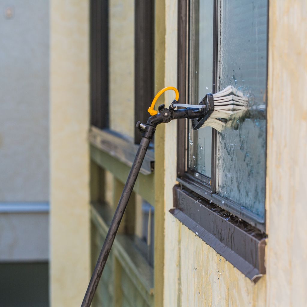 Window cleaning in Odessa, Texas. A water-fed pole brush scrubs a window on a tan building. The dark-framed window has water droplets running down the glass.
