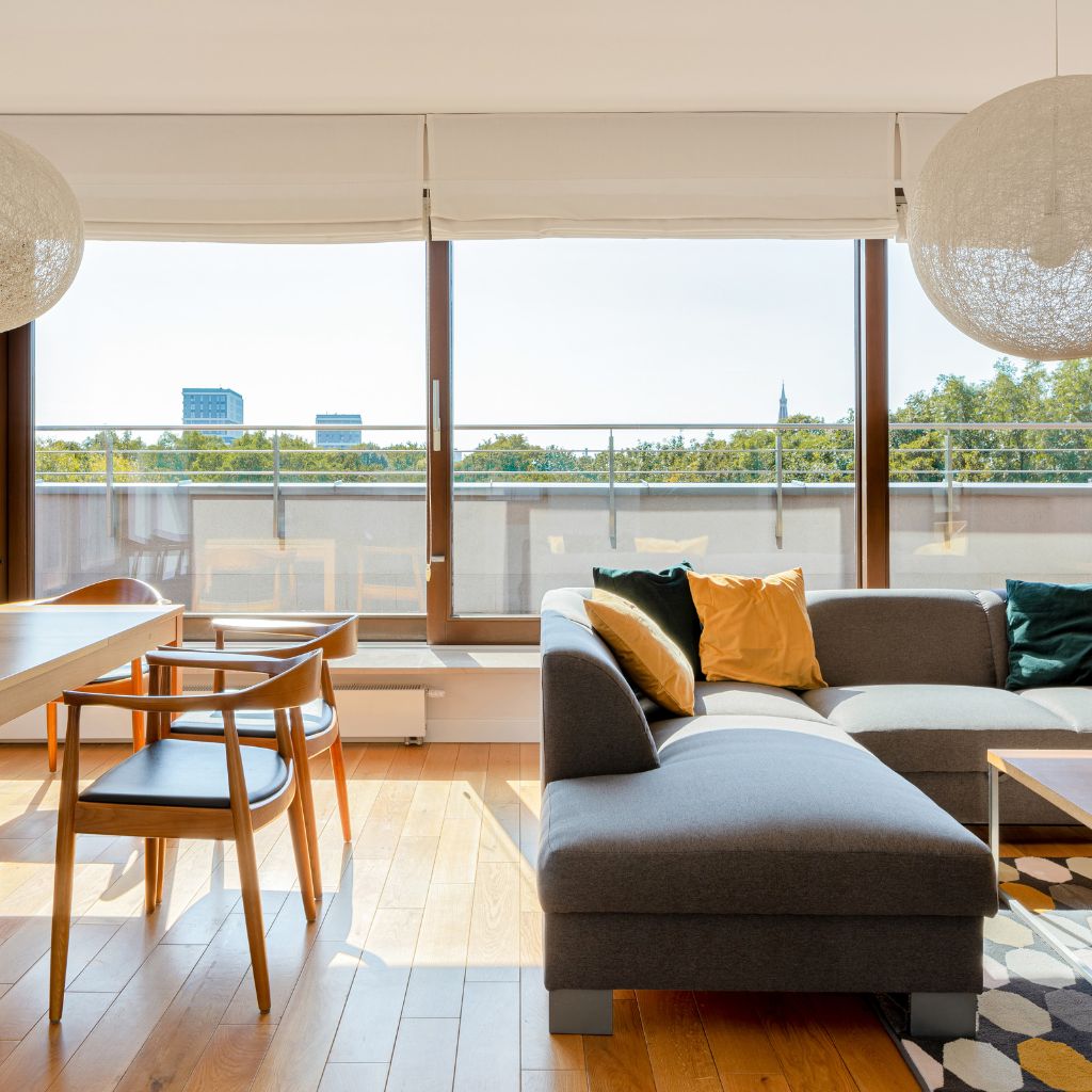 Sunlit modern living room with a gray sectional sofa adorned with yellow and green pillows, a wooden dining table and chairs, and large windows offering a view of Odessa, Texas. Window cleaning in Odessa, Texas keeps this room bright.