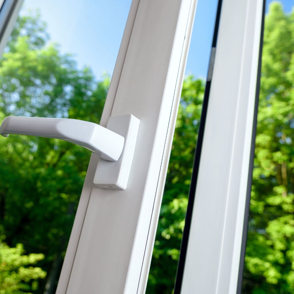 Close-up of a white window frame with a handle, showcasing the view of green trees and blue sky outside. A reminder of the fresh air and clean windows you can get with Atlas Scrubbed window cleaning in Midland, Texas.