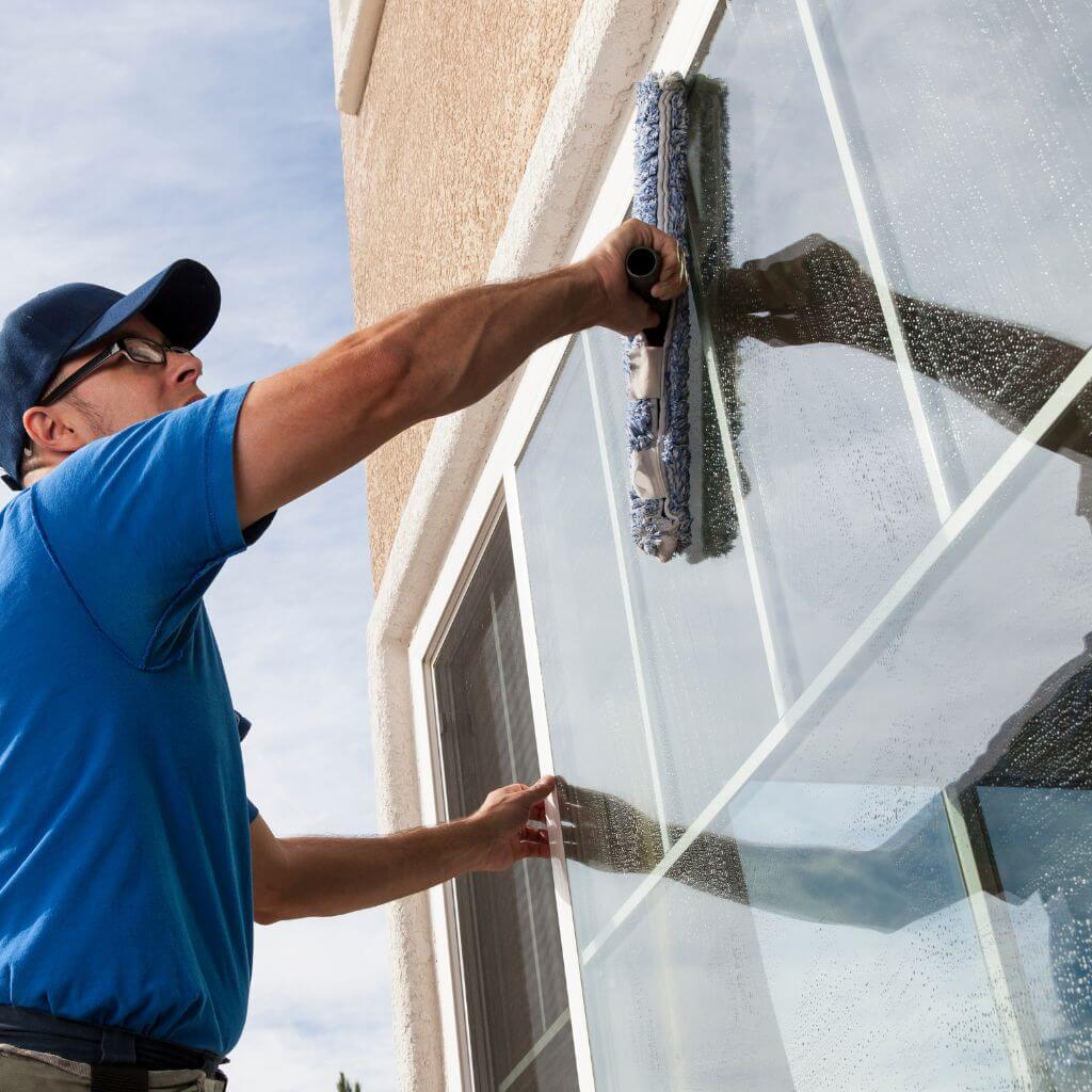 Window cleaner in Lubbock, Texas uses a scrubber to wash a large window. The Atlas Scrubbed professional wears a blue shirt and cap while working outdoors on a sunny day.