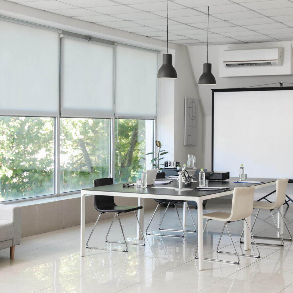 Bright, modern conference room with large windows covered by white blinds. A black table is surrounded by chairs, ready for a meeting. Sunlight streams through the window, highlighting the clean, reflective floor.