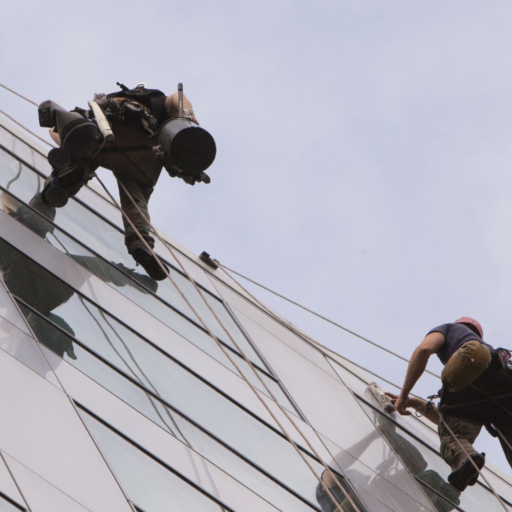 Two window cleaners suspended on ropes meticulously clean the exterior glass of a high-rise building. This image captures the skilled work of window cleaning in San Angelo, Texas, as performed by Atlas Scrubbed.