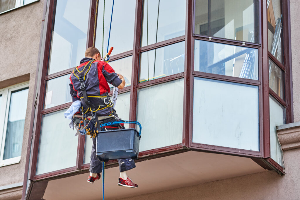 Midland window cleaning: A worker suspended by ropes cleans windows on a multi-story building, using a bucket and squeegee to ensure a clear view.