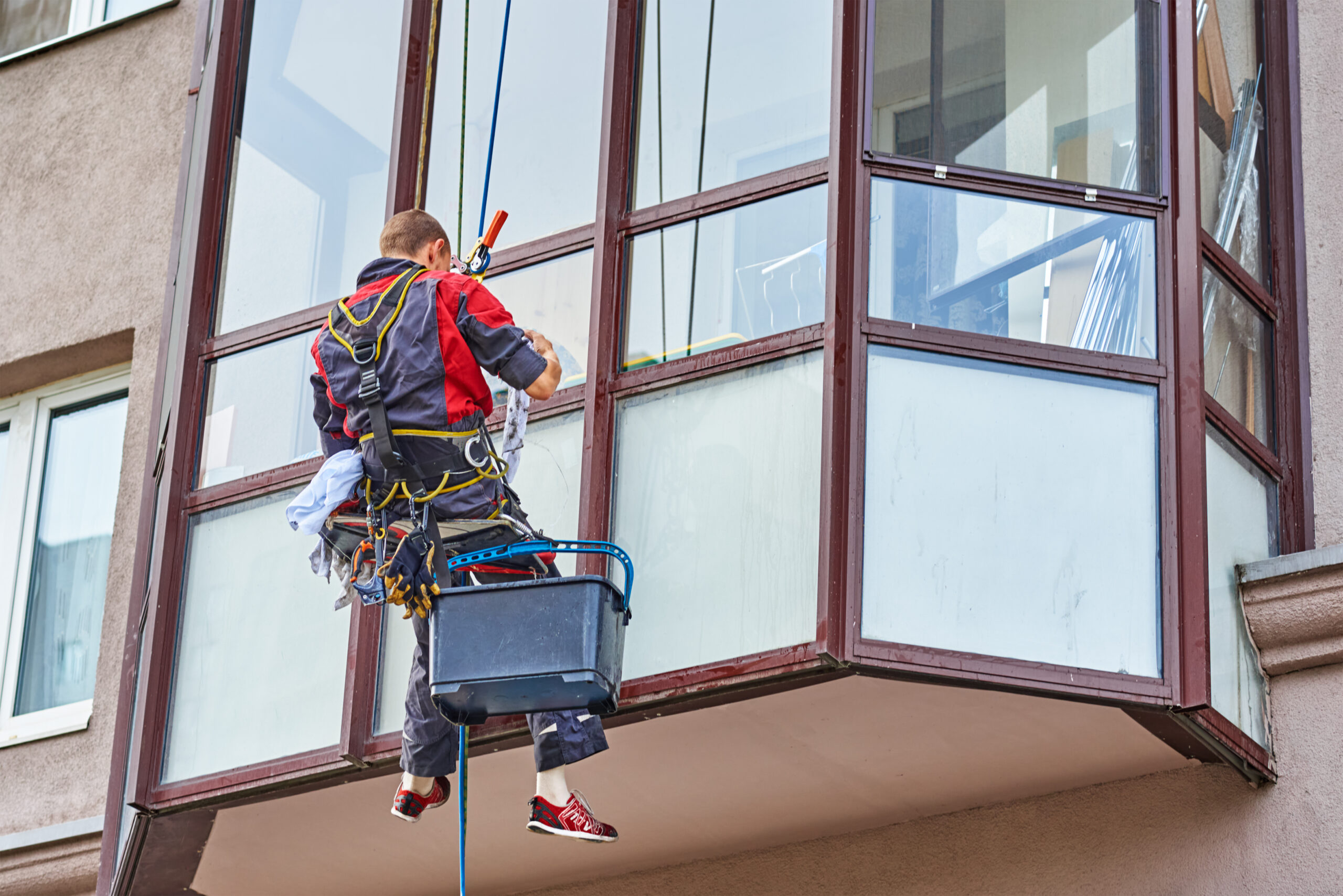 Midland window cleaning: A worker suspended by ropes cleans windows on a multi-story building, using a bucket and squeegee to ensure a clear view.
